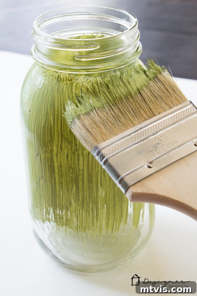 Craft Your Own Custom Mason Jars 3 Step-by-step image showing a mason jar with a vinyl stencil applied and the first coat of chalk paint being carefully brushed over the design.