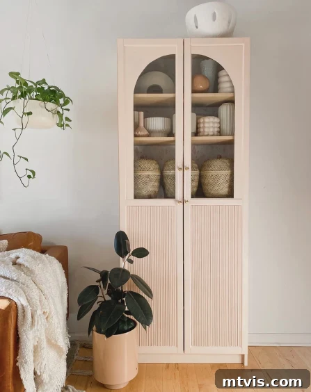 50 IKEA Hacks You Won't Believe Are IKEA 16 A tall, elegant cabinet featuring arched fluted doors, positioned beside lush potted plants, a light leather chair, and an off-white blanket. This sophisticated DIY IKEA hack seamlessly combines Billy bookcases with IKEA Oxberg cabinet doors, creating a custom and inviting aesthetic.