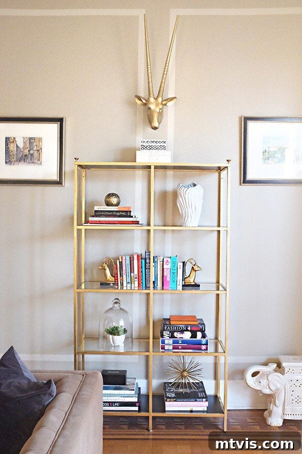 50 IKEA Hacks You Won't Believe Are IKEA 22 A luxurious gold storage rack, ingeniously crafted from an IKEA Vittsjo unit, stylishly holds an assortment of books and decorative items. Above the open shelves, a striking gold horned animal head serves as a focal point, flanked by elegantly framed pictures on either side, creating a glamorous display.