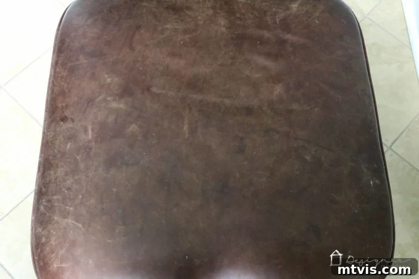 Close-up of a severely damaged leather ottoman, showing deep scratches, scuffs, and discolored marks.