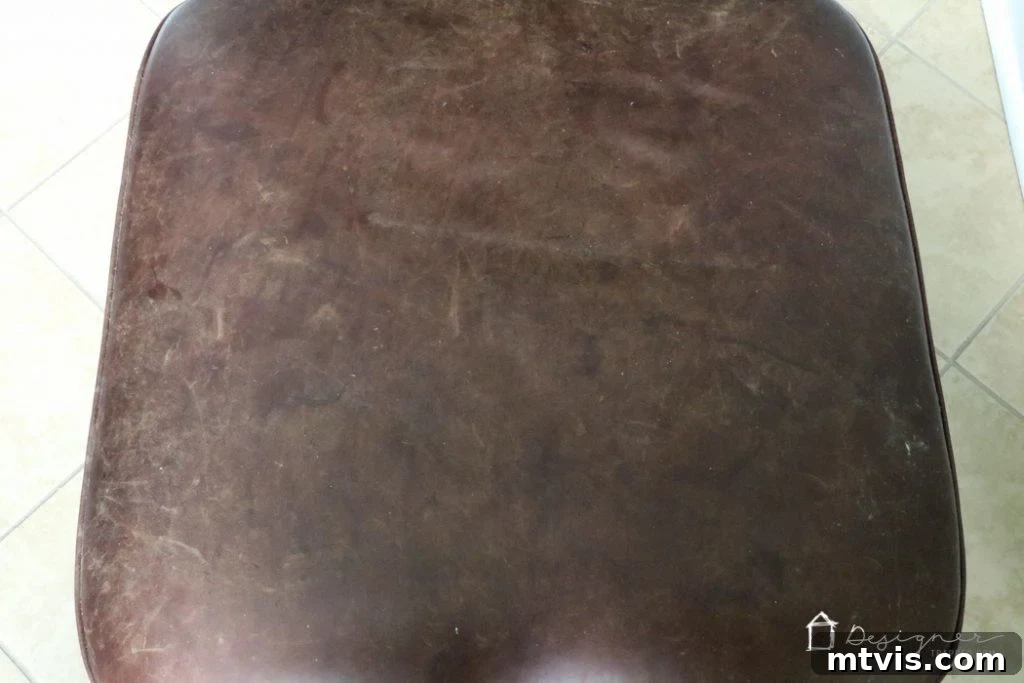 Before restoration: A damaged leather ottoman with noticeable scratches and wear.