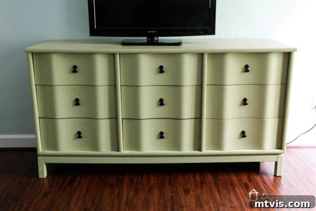 A light green-yellow dresser with curved drawer fronts stands against a light blue wall on a hardwood floor, topped with a flat screen TV. This sturdy piece of furniture is awaiting its stunning transformation through painting.