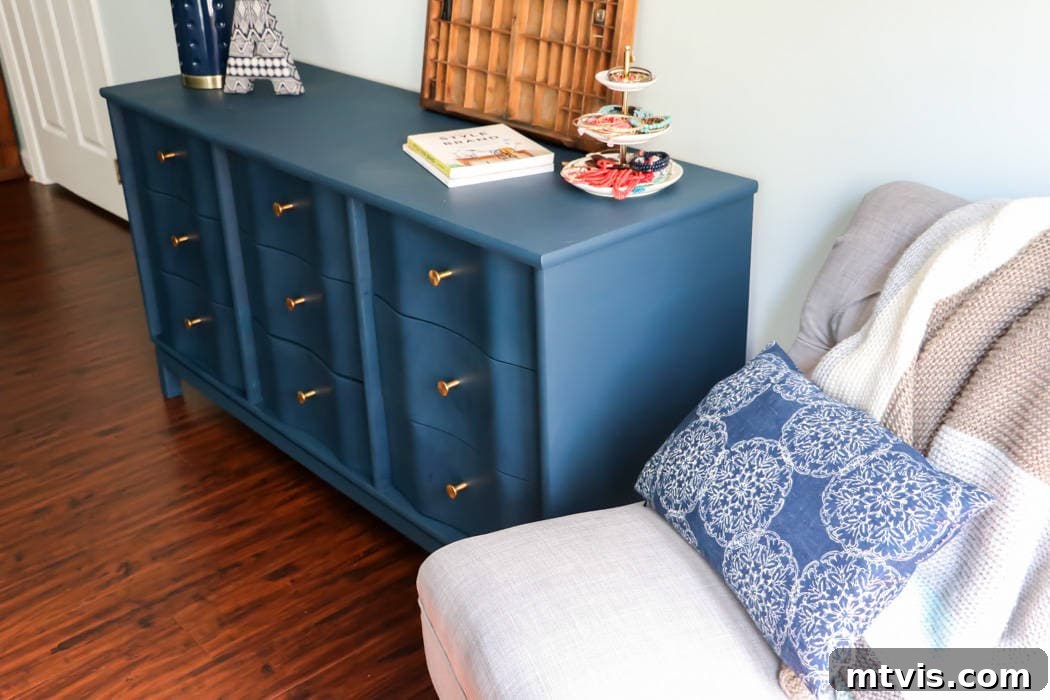 An angled perspective of a navy-blue painted wood dresser with striking gold knobs, decorated with a lamp, books, a three-tiered tea and cake stand, and other items on its top surface. To the right stands a cozy gray armchair featuring a blue patterned pillow and a soft knit throw. The subtle house outline logo and website, 'DesignerTrapped.com,' is in the lower corner. This dresser exemplifies expert techniques on how to paint wood furniture.