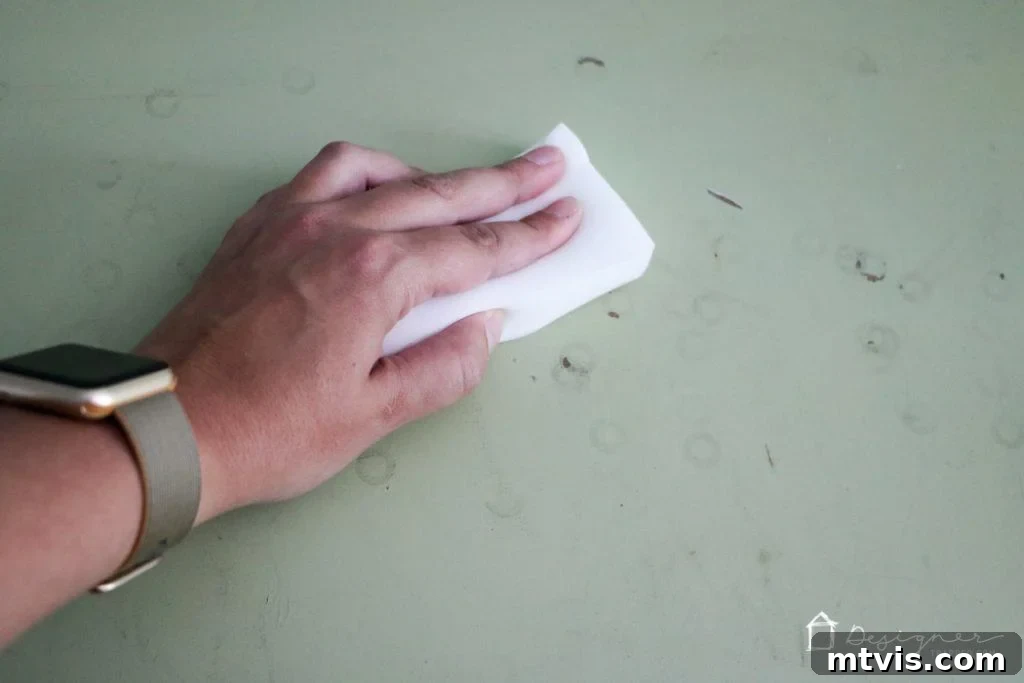 A hand wearing a green and gold watch actively uses a Mr. Clean Magic Eraser to scrub away dirt and stains from the top of an old green dresser, vividly demonstrating the thorough cleaning process before painting.