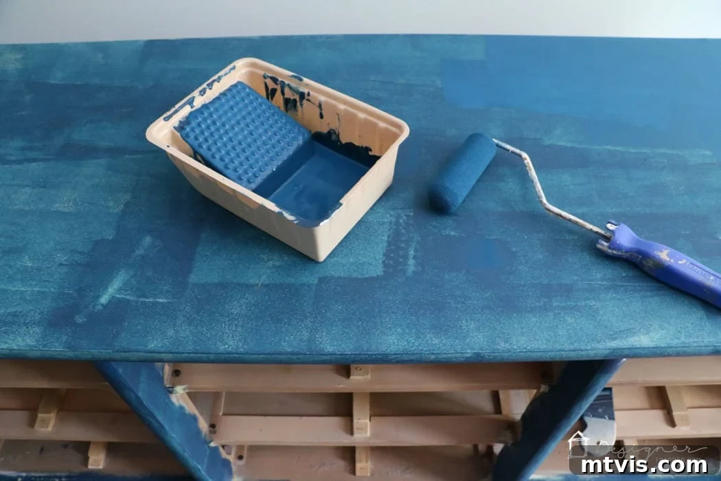 An overhead view captures a dresser being painted a deep blue. A paint tray and roller are positioned on top, with empty shelves visible below. The first layer of blue paint has been applied to the top and shelf supports, illustrating the initial stage of how to paint wood furniture.