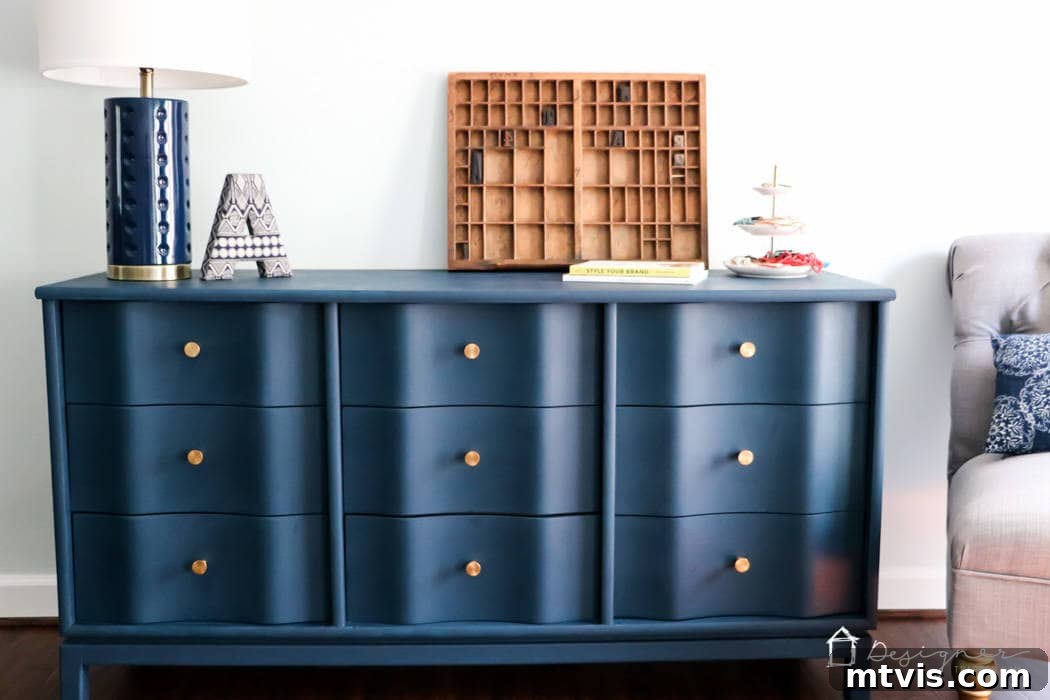 A bold navy blue dresser featuring gold knobs and elegant wavy drawer fronts boasts a smooth, pristine finish. Modern decor items and a stack of books adorn its top, while a stylish gray chair sits to its right. This successful DIY makeover stands as a prime example of how to paint wood furniture to achieve long-lasting, beautiful results.