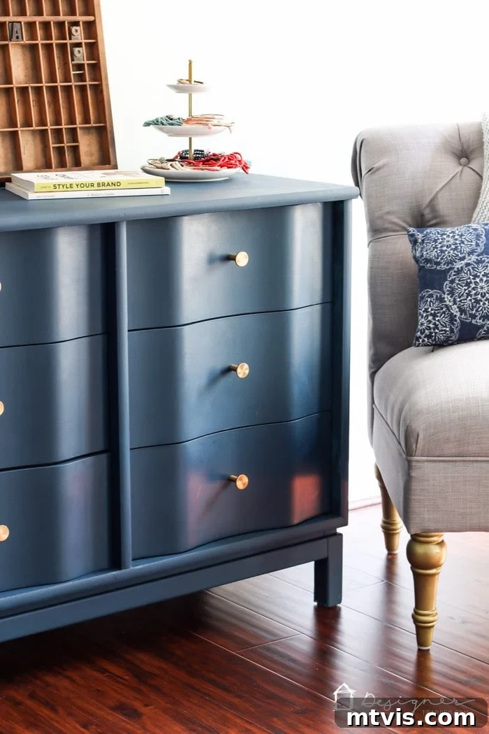A close-up shot of a navy blue painted wood dresser, adorned with elegant gold knobs. It is styled with a tiered tray of accessories, a stack of books, and a vintage letterpress, placed beside a comfortable tufted gray armchair. This polished example showcases how to paint wood furniture for a sophisticated, modern makeover. The text in the lower right corner says, 'DesignerTrapped.com' with a house logo.