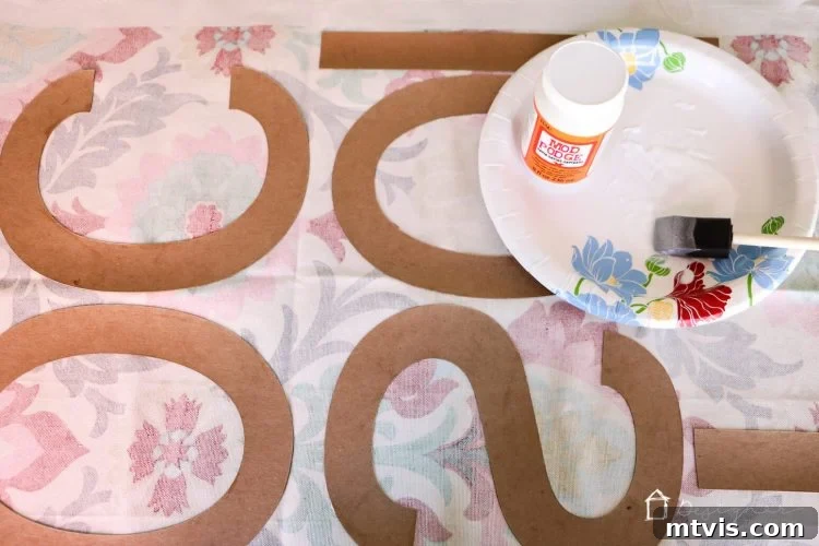 Applying Mod Podge to chipboard letters and pressing onto fabric to create personalized, textured wall decor.