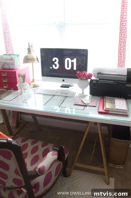 Smart IKEA Desk Upgrades on a Budget 6 A stylish IKEA desk hack features a repurposed door placed on gold sawhorse legs, topped with glass, along with other office essentials such as a computer monitor, printer, and pink accessories. "www.dwellingsbydevore.com" is printed in the corner.