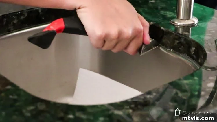 A hand using a caulk removal tool to scrape away old, discolored caulk from around a sink edge. Detailed caulk removal process.