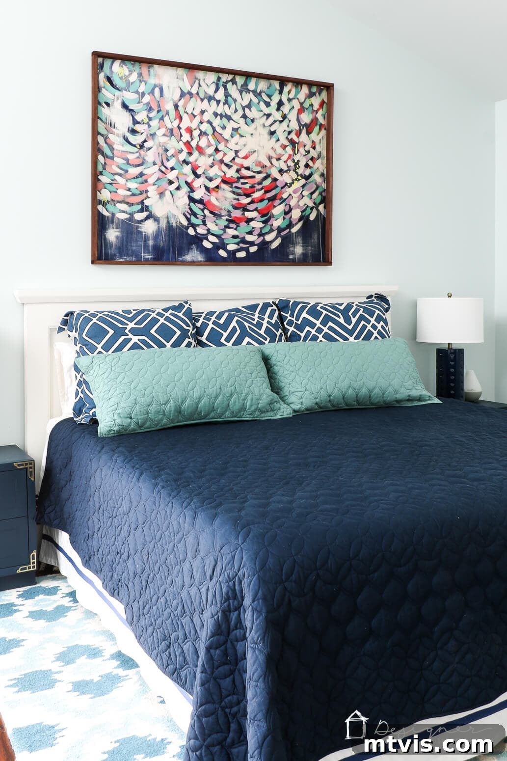 Build Custom Frames for Your Oversized Art 11 A large abstract art print, beautifully framed in a handmade picture frame, hangs above a bed adorned with teal and blue pillows and a matching blue bedspread. A white headboard and a complementary white and blue lamp complete the serene bedroom decor.
