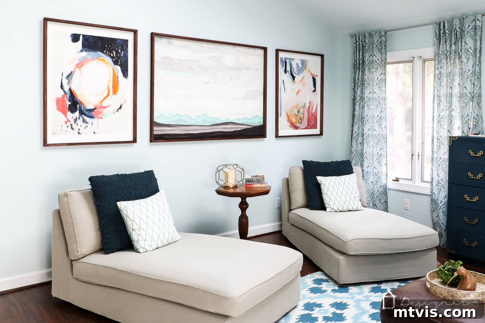 Build Custom Frames for Your Oversized Art 13 A serene living room features light blue walls adorned with three abstract art prints, each encased in custom wooden frames made using this DIY tutorial. The artwork is positioned above two elegant cream-colored chaise loungers, accented with navy and white pillows, with a round side table nestled between them.