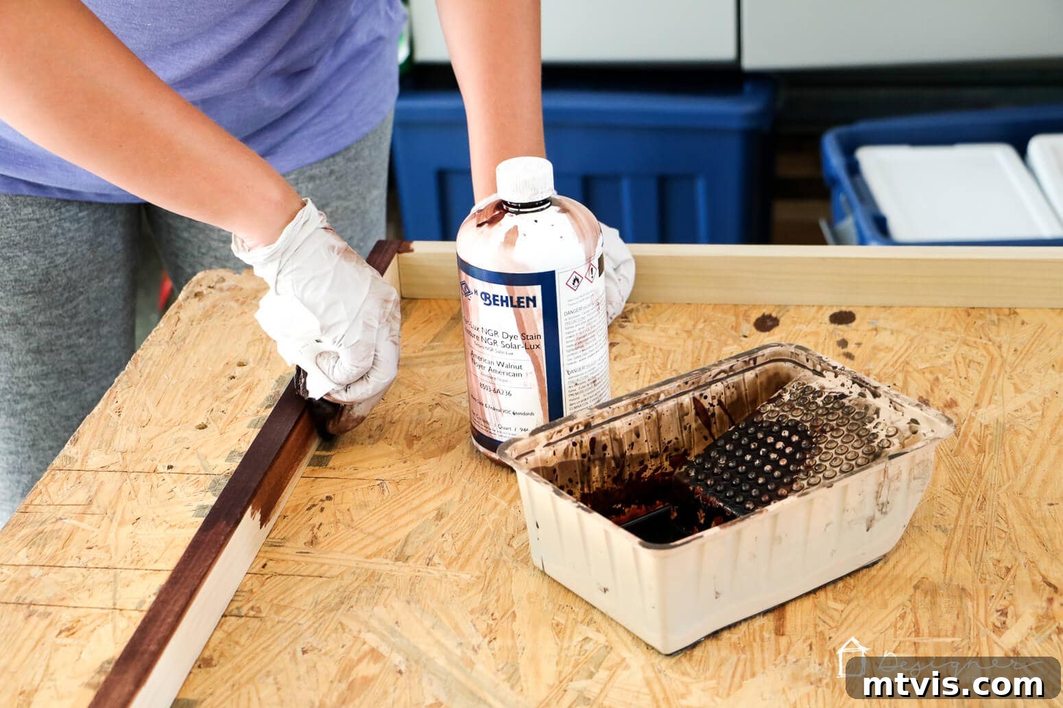 Build Custom Frames for Your Oversized Art 7 Tasha, wearing white gloves, applies a rich American Walnut dye stain to a DIY wooden frame, enhancing its natural grain. A jar of Behlen Solar Lux Dye Stain and a paint tray are visible on the plywood workbench, illustrating the staining process.