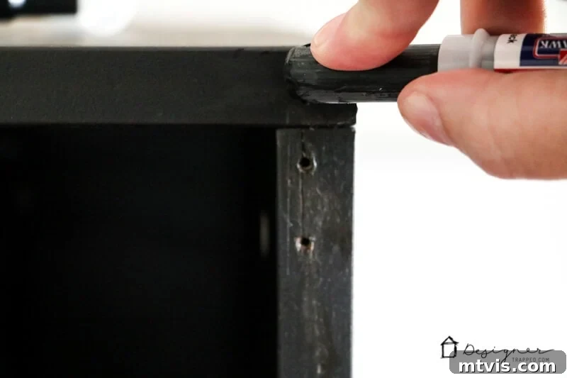 Seamlessly Repair Furniture to Match IKEA Hues 7 A zoomed-in view of a hand applying a furniture repair stick to mail holes in the back of IKEA furniture. The words, "DesignerTrapped.com" are in the bottom corner next to a simple house logo.