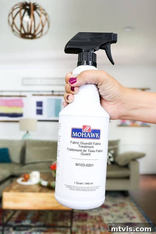 A woman's hand securely grips a white spray bottle of Mohawk Fabric Guard, prominently displaying the product label, which is recommended as an effective fabric protection solution.