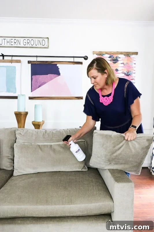 Tasha, with focused attention, applies Mohawk Fabric Guard to her light-colored sectional couch. She is in a bright, inviting room featuring a vibrant rod of colorful artwork displayed on the wall behind the sofa, adding a touch of artistic flair to the space.