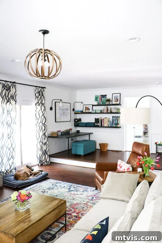 Bright, modern living room with a cozy reading nook and home office space, with a sectional sofa, leather chair, wood coffee table with fresh flowers, floating bookshelves, and patterned curtains. A dog lounges by the sliding glass doors.