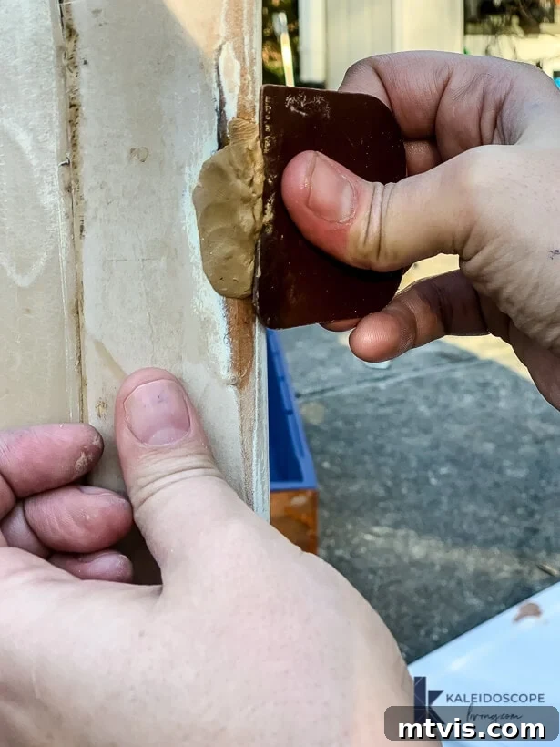 Hands use a scraper to smooth epoxy wood filler putty onto the damage wood while outside. 