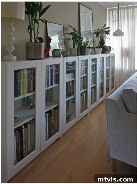 IKEA Smart Hacks: Custom Built-Ins and Bookshelf Upgrades 4 A row of white, medium height IKEA bookcases with glass doors. Plants, photographs and a lamp rest on top. A couch is visible in front of bookcases.