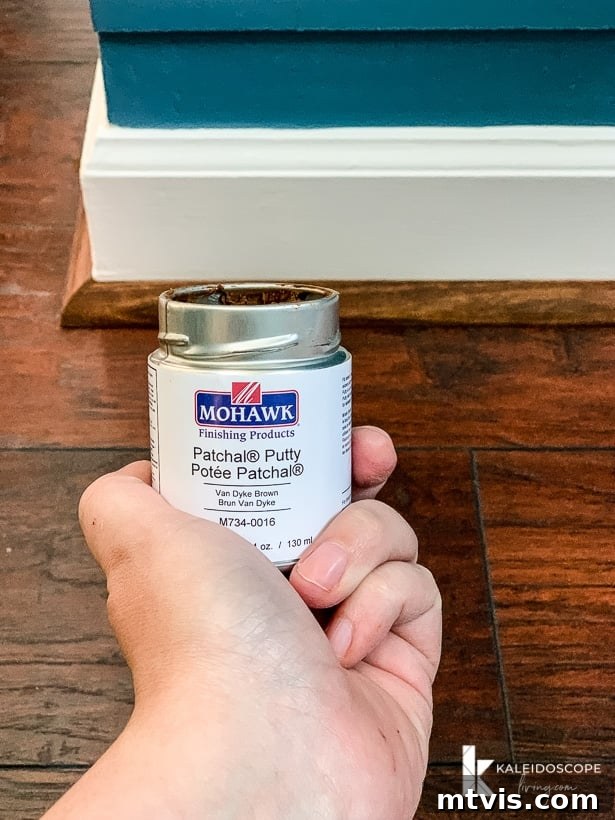 DIY Quarter Round Molding Installation 10 A hand holds a jar of Mohawk Patchal Putty in front of quarter round molding, a white baseboard, and a blue wall.
