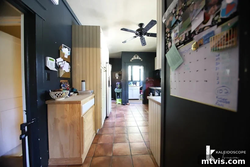 DIY Kitchen Transformation Under One Thousand 3 An outdated kitchen before a budget-friendly kitchen makeover removed the dark cave-like walls and trim. A family calendar hangs on the side of a refrigerator and brown tiles line the floor and hallway into the kitchen,