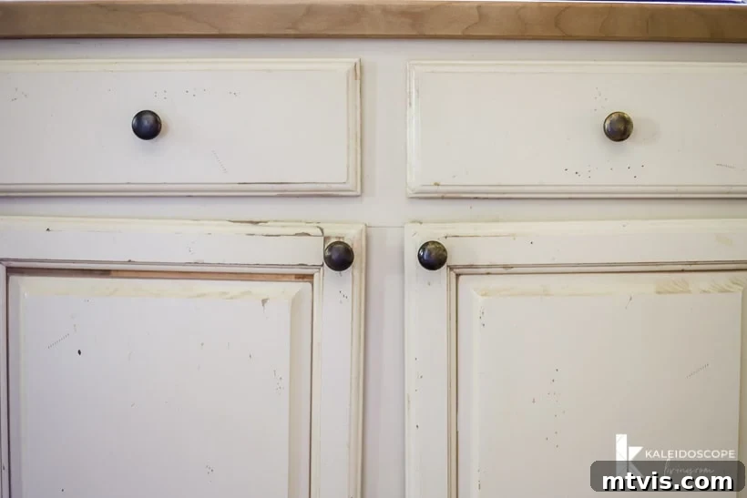 DIY Kitchen Transformation Under One Thousand 6 Worn out painted kitchen cabinets with uneven hanging doors, dirt, and paint scratches. The corner text reads, "KaleidoscopeLiving.com."