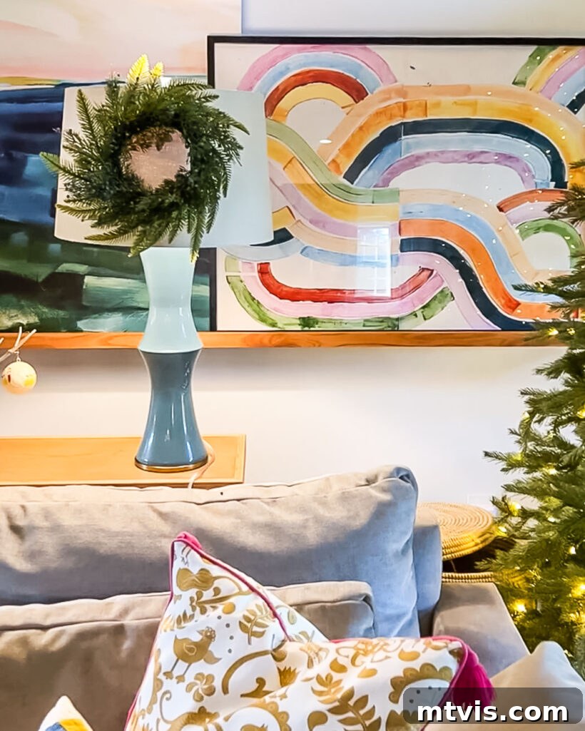 A mini wreath hanging on a lampshade in colorful family room decorated for Christmas, with the top of a gray couch and framed artwork on the wall. 