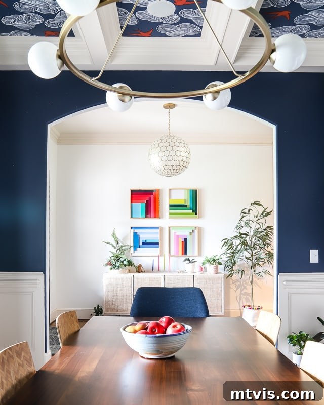 Step Inside Our Navy Blue Dining Room 7 A beautifully transformed dining room, painted in a deep Gale Force blue, showcases a stunning wallpaper and painted coffered ceiling. The view extends into a bright white foyer adorned with four colorful square art pieces on the wall, creating a vibrant contrast.