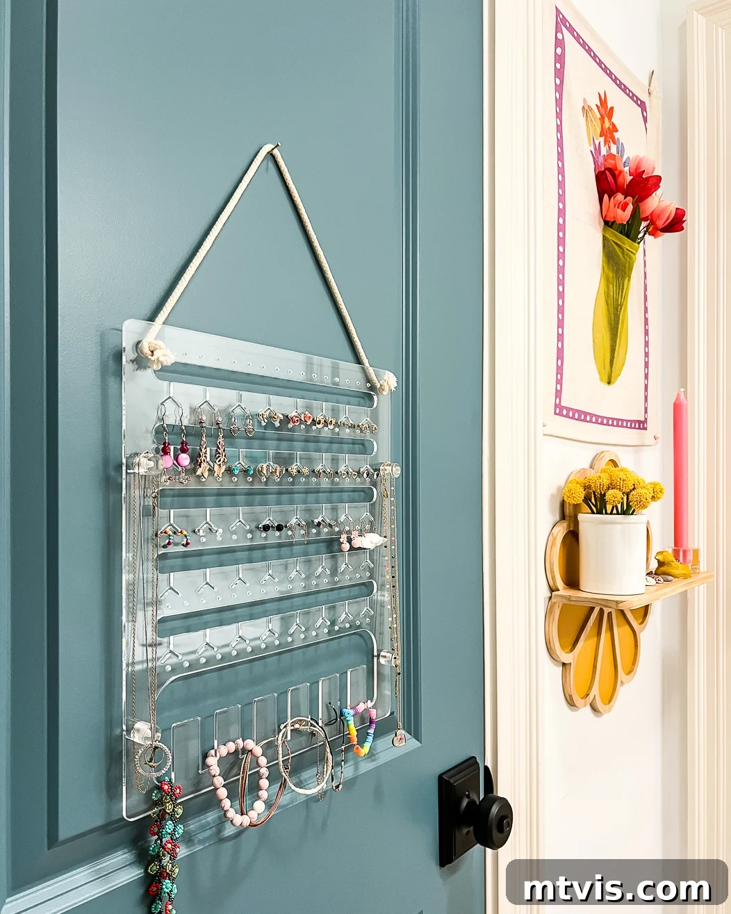 A clear acrylic hanging jewelry organizer in girl's bathroom on the back of a green-blue door with yellow flowers on the wall to the side. 