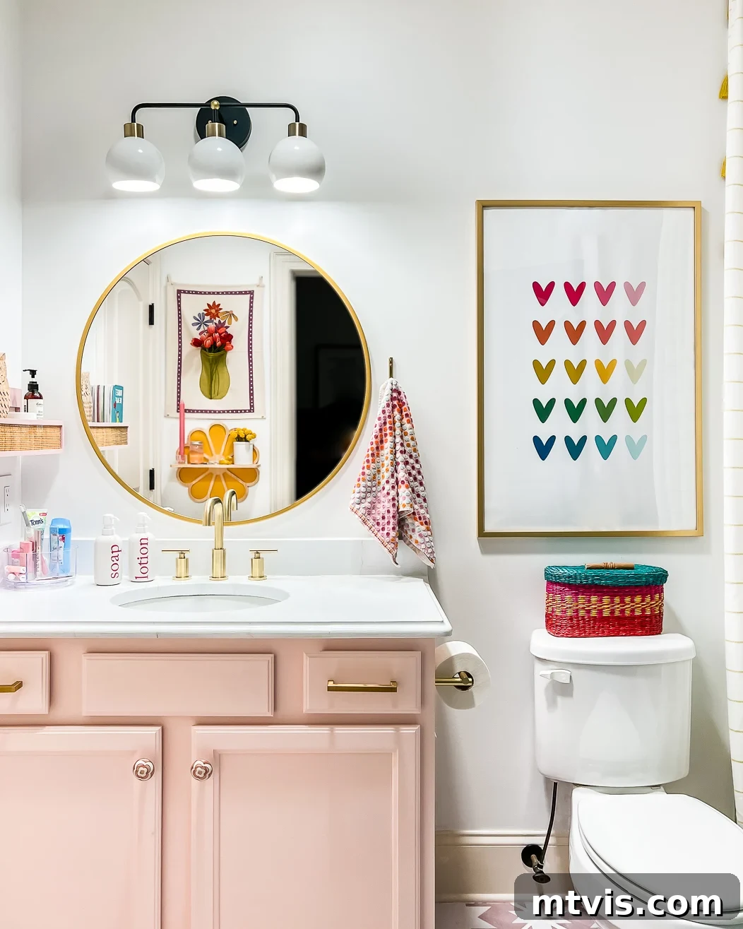 A bathroom makeover on a budget includes a round gold mirror, white walls with pink cabinets, rainbow heart artwork. toilet, and flower artwork reflected in the mirror. 