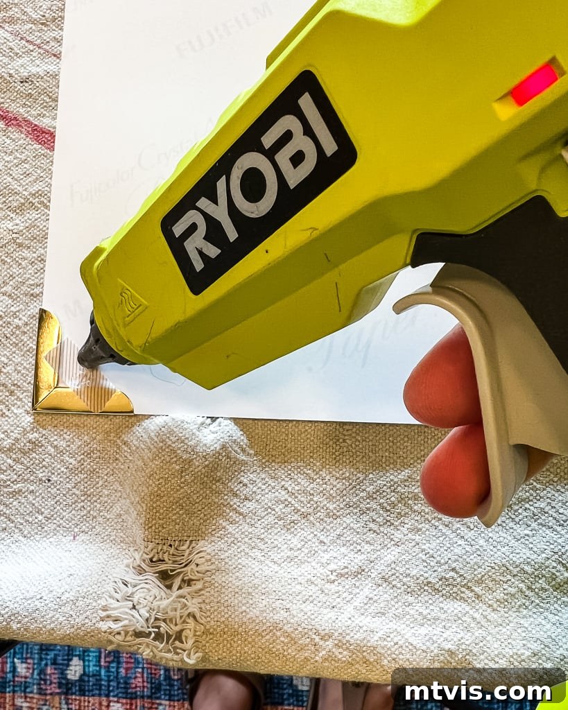 A hot glue gun is used to apply small dots of glue to the back corners of a photo, preparing it for attachment to the decorative mat.