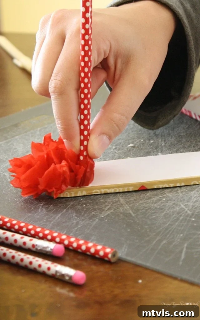 Valentines Door Decor DIY 13 A hand using a pencil to press a red tissue paper square onto a glue-covered foam core letter, creating a fluffy texture.