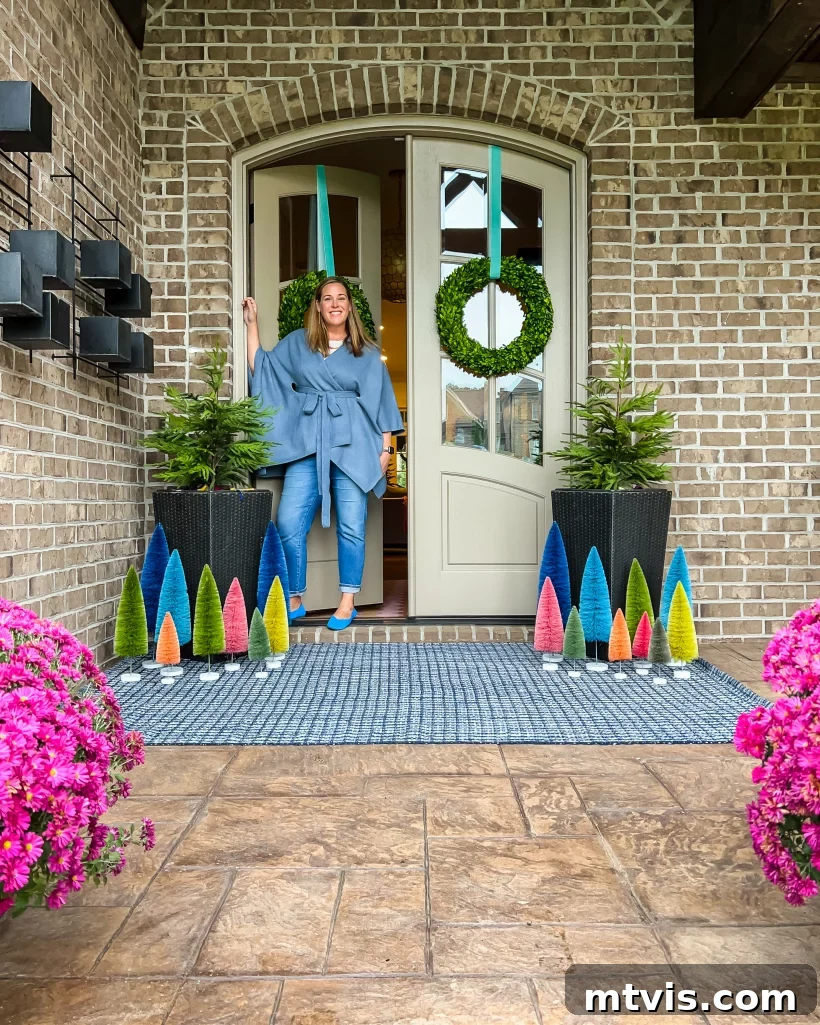 Festive Front Porch Christmas Magic 3 Tasha Agruso stands on her front porch among Colorful front porch Christmas decorations, including vibrant colorful bottle brush trees of various sizes, mini evergreen trees in large planters and wreaths on the front door.