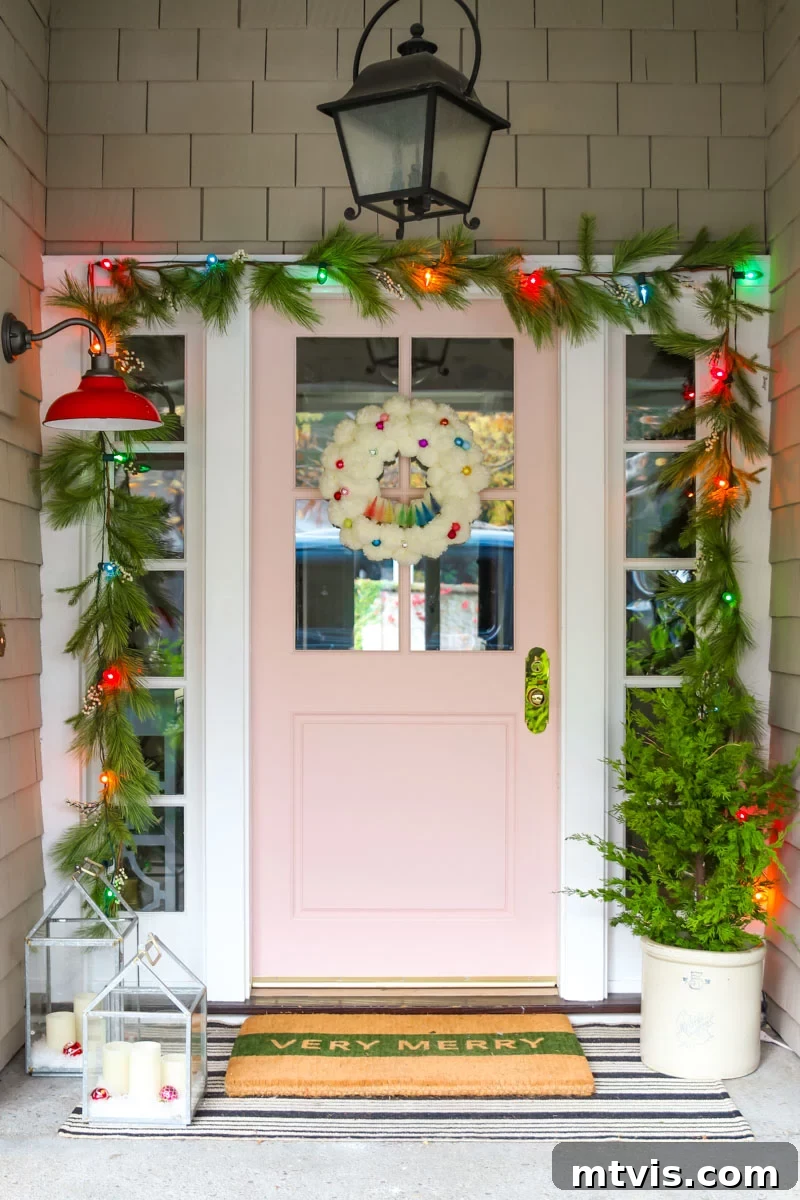 Festive Front Porch Christmas Magic 7 A blush-colored door in the middle of large brick walls has a sparse garland with multicolored lights framing the door, and other front porch Christmas decorations including a mini evergreen in a white plants, and candles in glass cases. These front porch Christmas decorations were done by Modern Glam.