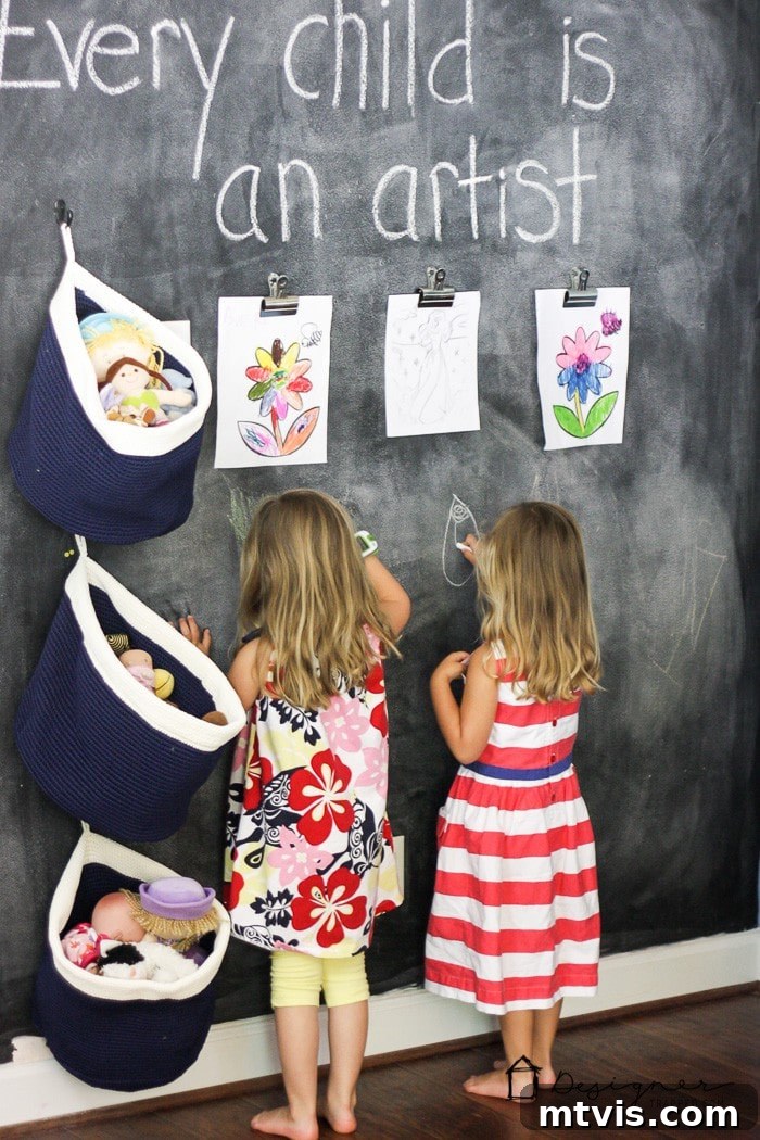 Playful and Chic Playroom Design Guide 22 Two young girls write on a floor to ceiling chalk wall. Children's art work hangs above them, bins full of staffed animals hang on the side, and the top of the wall reads,