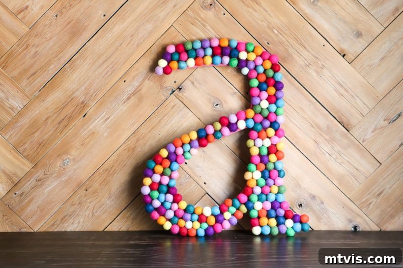 23 Ingenious DIY Wall Art Projects 19 A large DIY rainbow felt ball letter, vibrantly colorful, casually leaning against a textured wood wall, adding a playful and personal touch.
