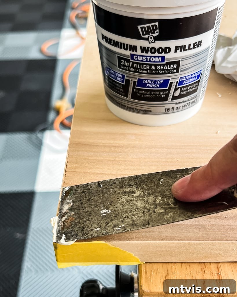 A putty knife applying wood filler to smooth out imperfections on a plywood surface, preparing it for painting in the DIY dog crate construction.
