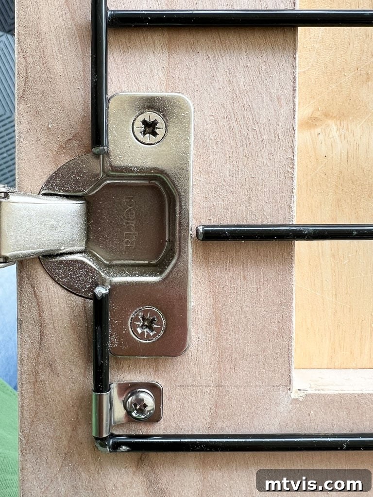 Close-up photograph showing a concealed door hinge installed within the door frame of a wooden DIY dog crate, with wire clamps securing the internal panel.