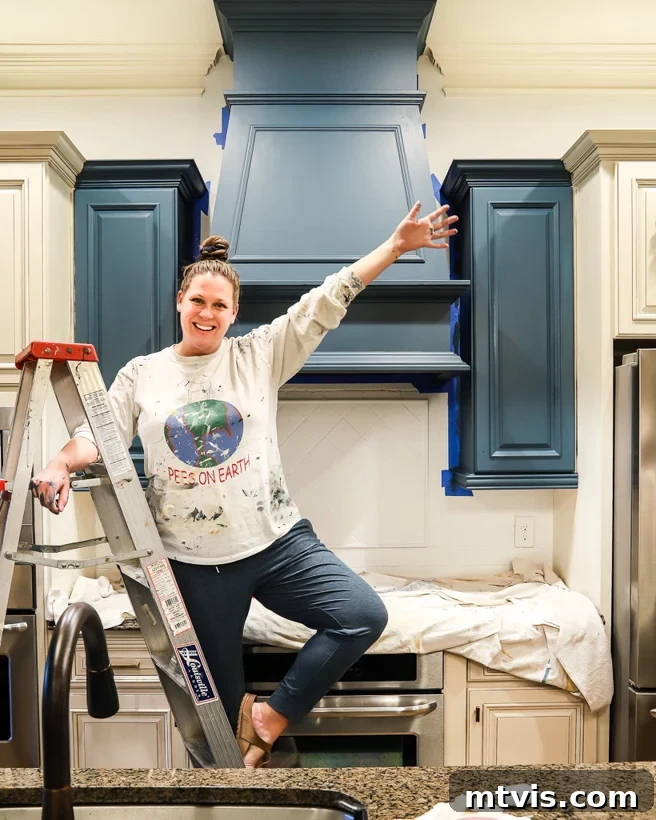 My Triple-Tested Method for Perfectly Painted Kitchen Cabinets 2 photo of Tasha Agruso of Kaleidoscope Living standing on a ladder in painting clothes in front of partially painted kitchen cabinets, illustrating the DIY process