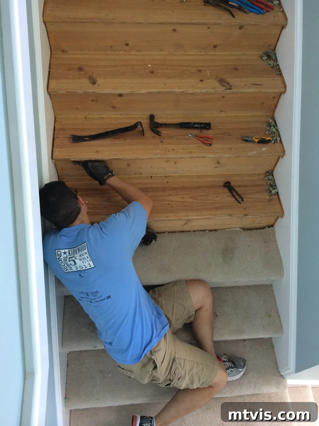 Man removing old carpet from stairs, revealing the plywood subfloor underneath, preparatory step for DIY hardwood stairs